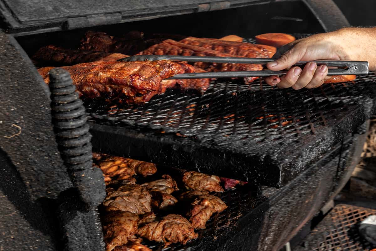Close-up of ribs being grilled on a smoky barbecue pit, with a hand using tongs to turn the meat.