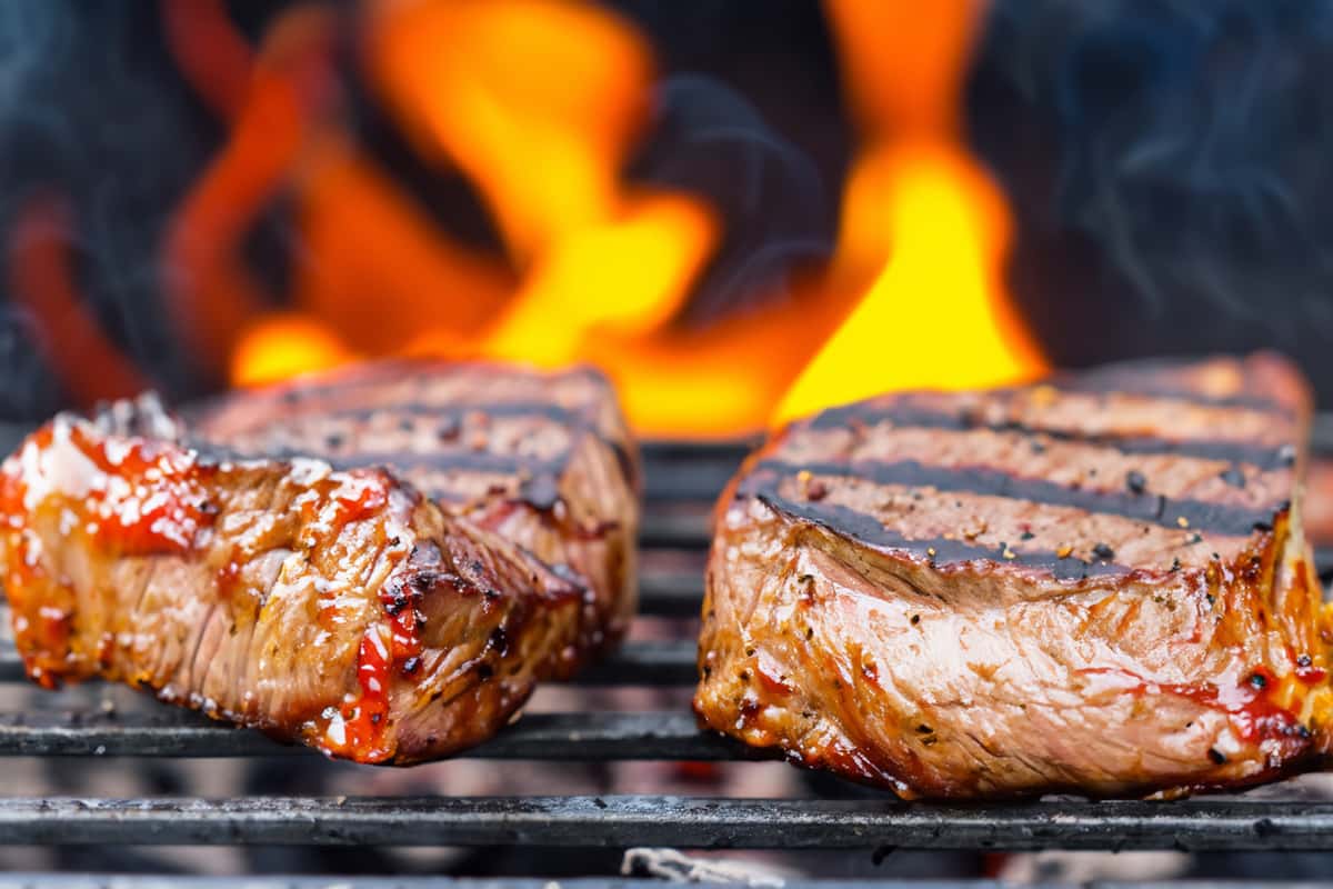 Juicy grilled steaks with seared grill marks cooking over open flames on a barbecue grill.