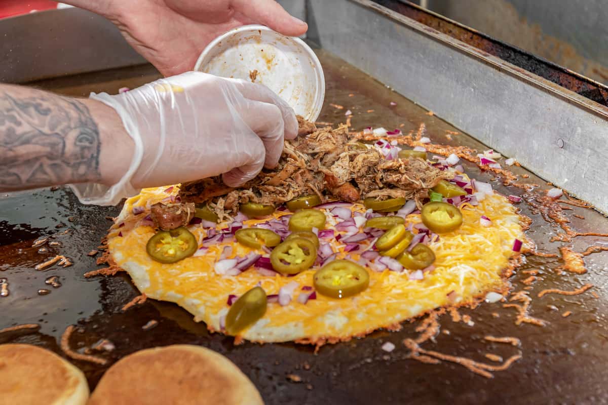 Cook preparing a cheesy quesadilla topped with pulled pork, jalapeños, and diced onions on a flat-top grill.