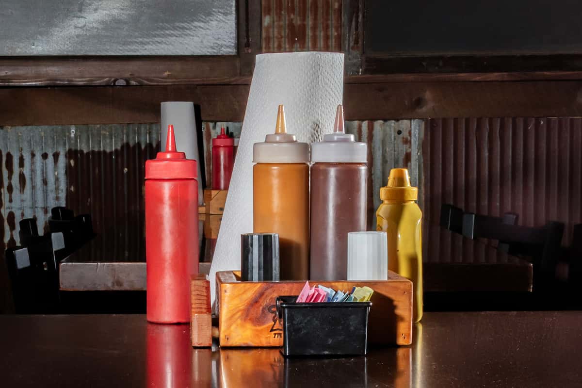 Restaurant table setup with condiment bottles for ketchup, mustard, and barbecue sauces, alongside napkins and sweetener packets.