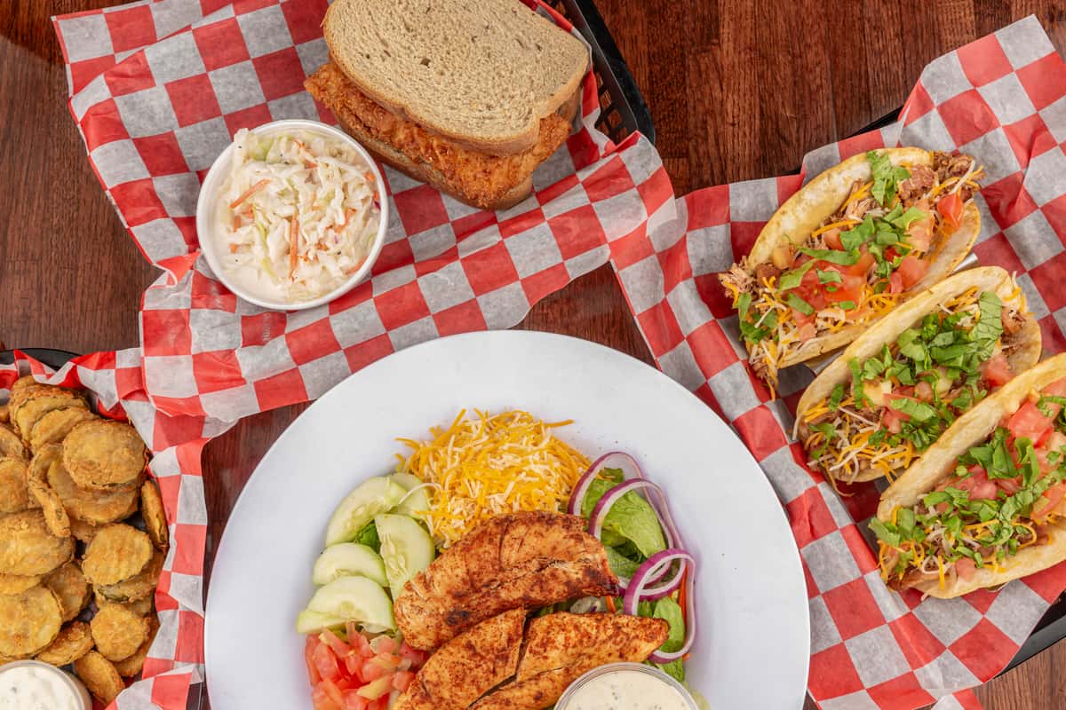 Assorted barbecue dishes including fried pickles, grilled chicken salad, pulled pork sandwich with coleslaw, and tacos with lettuce and tomato.