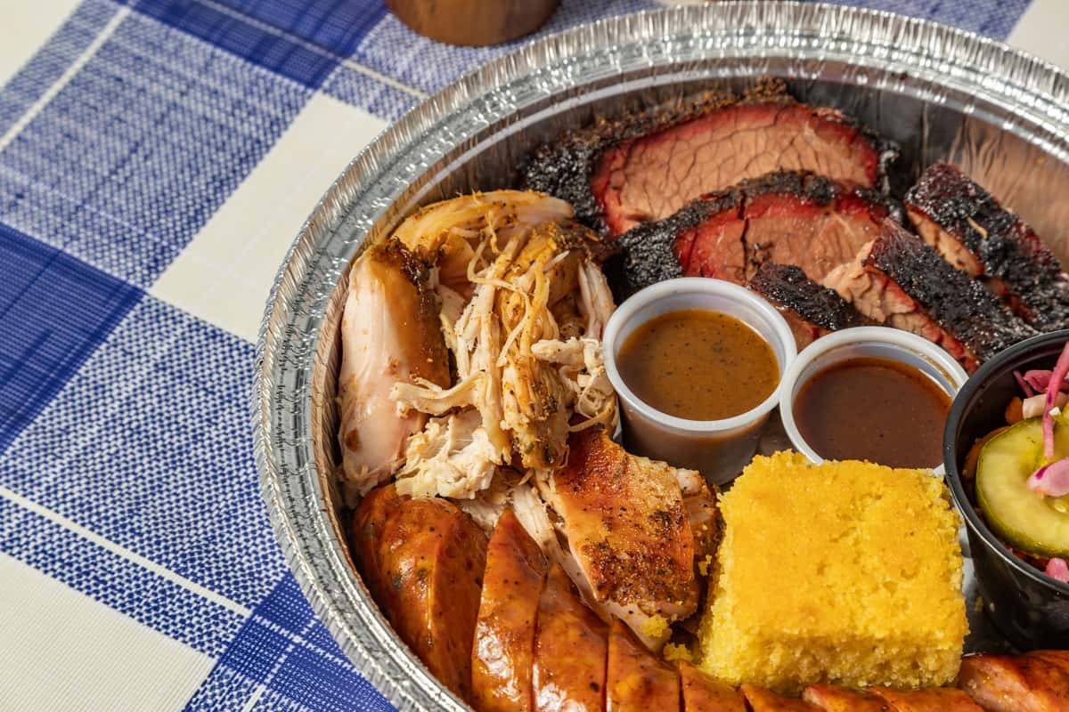 Barbecue combo platter in a foil tray featuring sliced smoked brisket with a dark bark, pulled chicken, grilled sausage links, a square of golden cornbread, barbecue sauces, and a side of pickles and slaw—perfect for Texas-style BBQ takeout or catering.