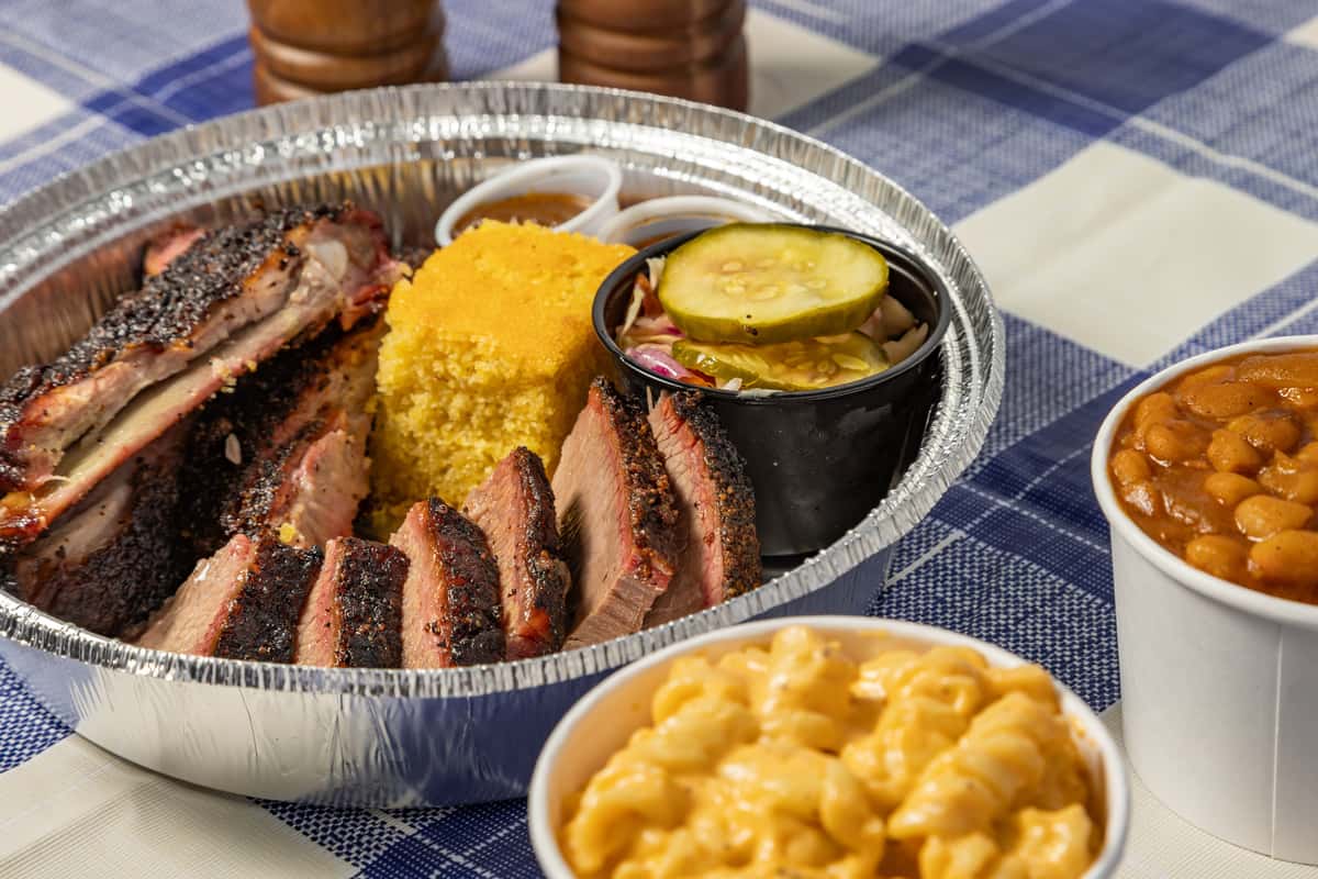 Barbecue sampler platter in a foil tray featuring smoky pork ribs, sliced brisket, cornbread, pickles, and slaw, served with a side of creamy mac and cheese and barbecue baked beans—ideal for Southern comfort food catering or takeout.