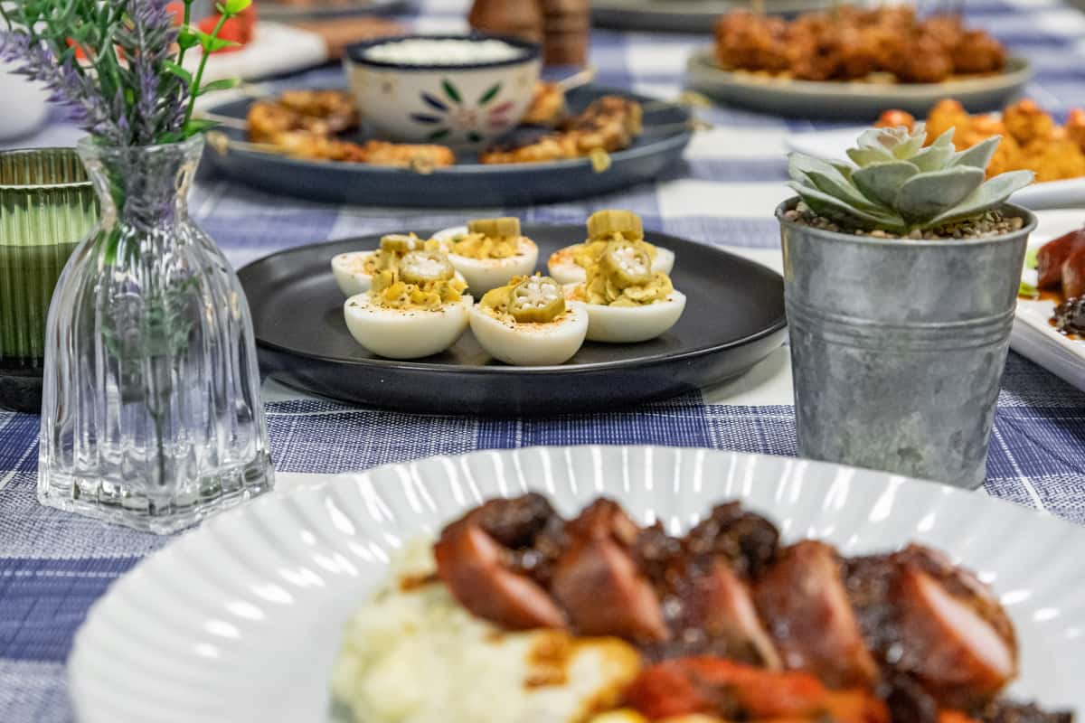 Close-up of Southern-style deviled eggs topped with pickled okra on a black plate, surrounded by assorted appetizers and floral decor atop a blue gingham tablecloth—perfect for a rustic brunch or homestyle catering event.