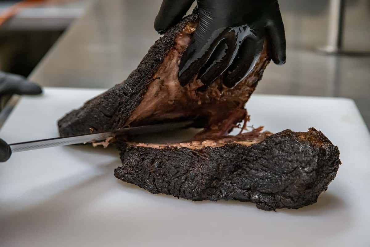 Close-up of a pitmaster slicing smoked beef brisket with a charred bark exterior and juicy interior, using a carving knife and wearing black gloves in a professional barbecue kitchen.