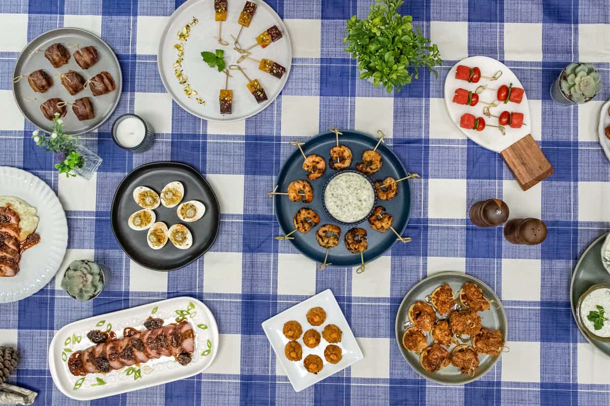 Flat lay of assorted gourmet appetizers arranged on a blue and white checkered tablecloth, featuring grilled shrimp skewers with herb dip, deviled eggs, bacon-wrapped bites, mini crab cakes, spicy fried chicken pieces, sliced pork tenderloin, and fresh watermelon skewers with tomatoes and mozzarella—perfect for a Southern-inspired brunch or upscale buffet spread.