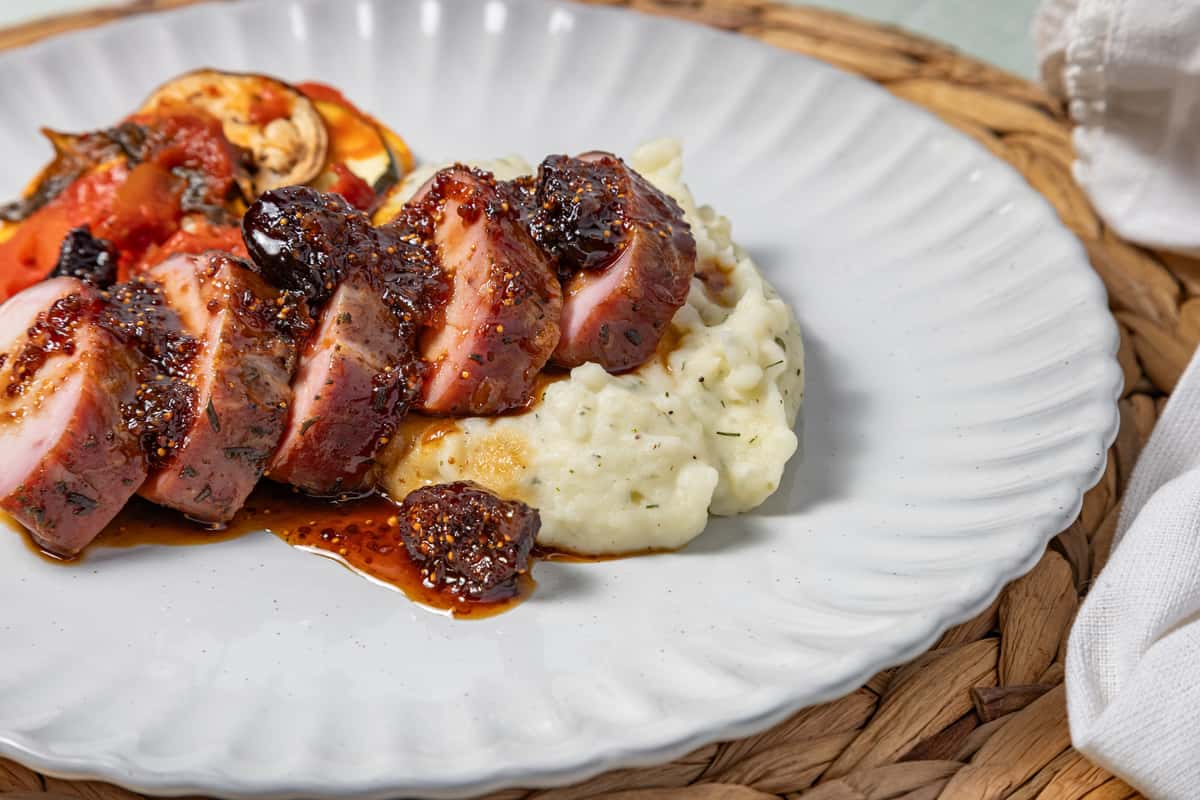 Sliced glazed pork tenderloin drizzled with a fig and herb reduction, served over creamy mashed potatoes with a side of roasted ratatouille-style vegetables, presented on a white scalloped plate with rustic table decor.