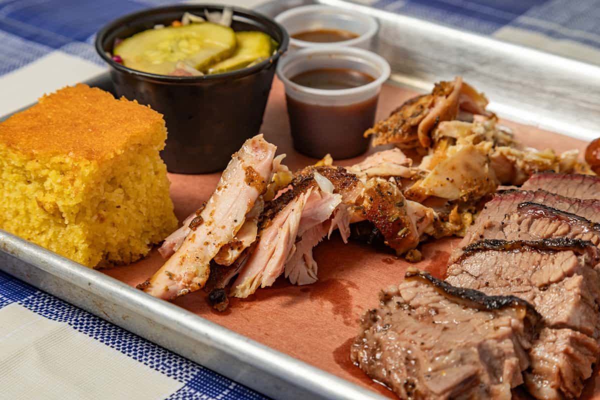 Texas-style barbecue platter featuring sliced brisket, pulled smoked chicken, a thick square of golden cornbread, dill pickles with coleslaw, and barbecue sauce served on a metal tray with butcher paper.