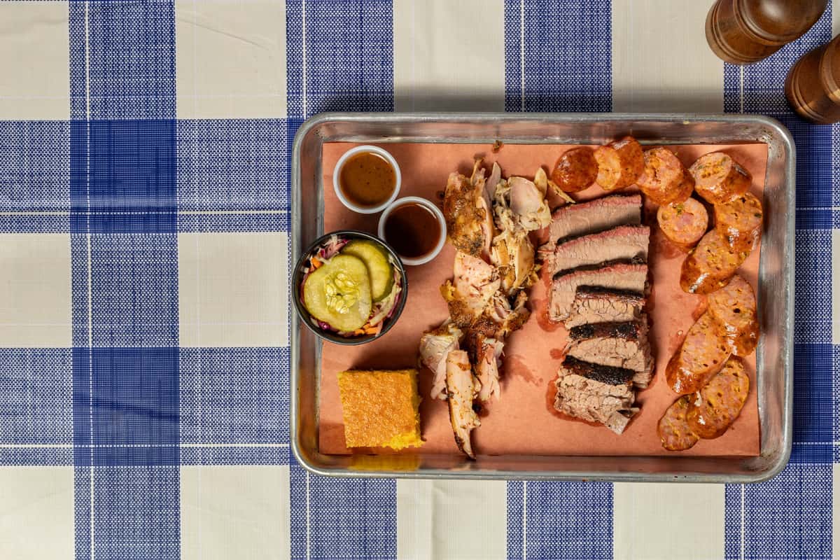Texas-style BBQ platter served on a metal tray lined with butcher paper, featuring sliced smoked brisket, juicy pulled chicken, spiced sausage links, cornbread, tangy pickles with slaw, and two house-made barbecue sauces—set atop a blue checkered tablecloth for a classic Southern dining experience.