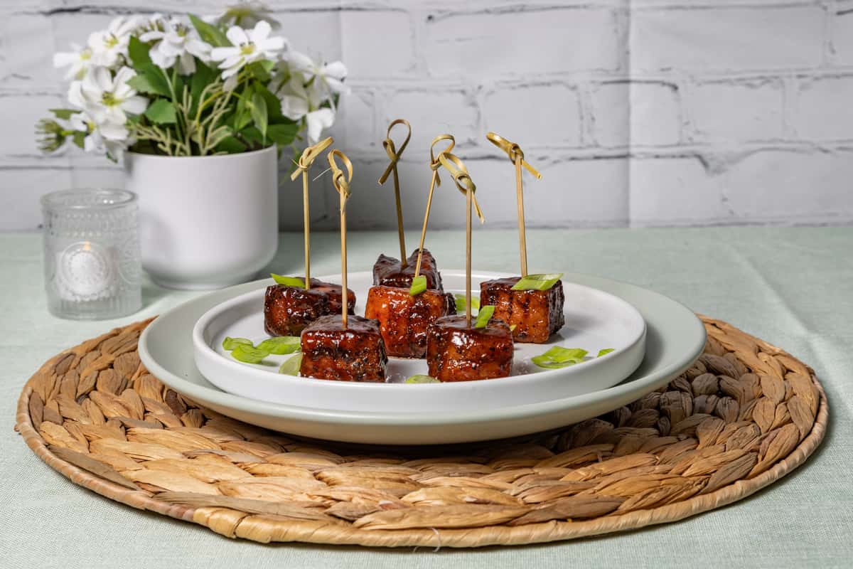Elegant appetizer plate featuring glazed tofu bites skewered with bamboo picks, garnished with fresh scallions, served on a minimalist white plate over a woven placemat with a soft floral background—perfect for a modern vegan or Asian-fusion restaurant setting.