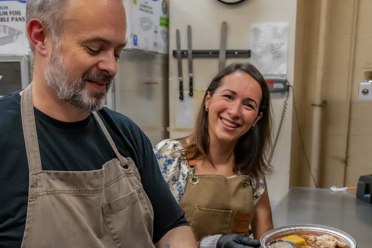Two smiling chefs in aprons preparing a to-go container with hearty barbecue dishes inside a commercial kitchen, featuring pulled meat, pickles, and a lemon wedge—capturing the warm, behind-the-scenes energy of a casual Southern-style restaurant