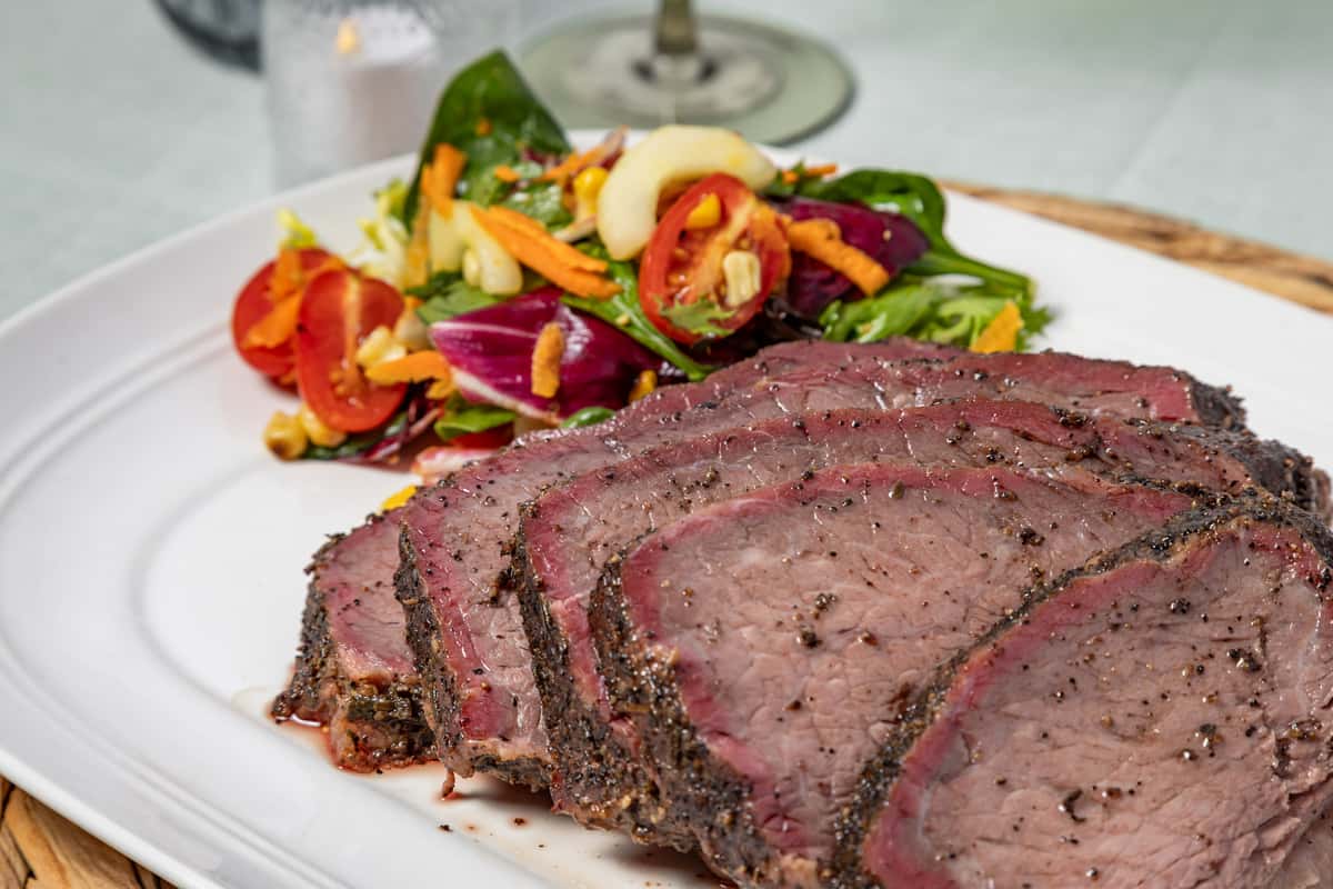 Sliced pepper-crusted roast beef served with a vibrant mixed green salad topped with cherry tomatoes, shredded carrots, and cashews, plated on a white dish in a fine dining setting.