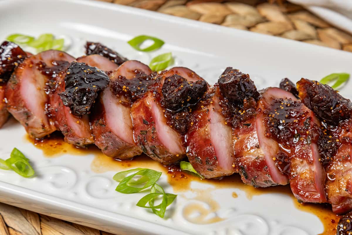 Close-up of glazed pork tenderloin medallions topped with a dark fig or cherry reduction, garnished with sliced scallions and served on a decorative white platter—perfect for elevated Southern cuisine or holiday mains.