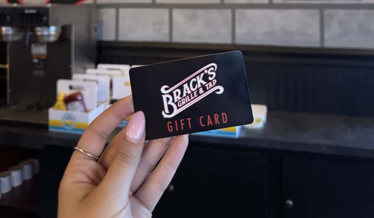 Brack's Grille & Tap gift card held in front of the restaurant's signature logo wall, perfect for dining, drinks, and special occasions.
