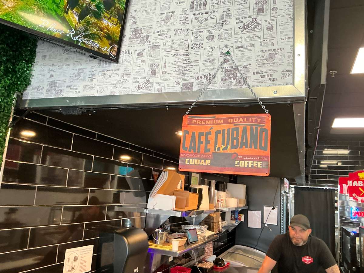 Here's a description of the image: A restaurant counter with a "Cafe Cubano" sign hanging above it, promoting Cuban coffee. The backdrop includes decorative, newspaper-style wallpaper, complementing the cafe's vintage Cuban theme.