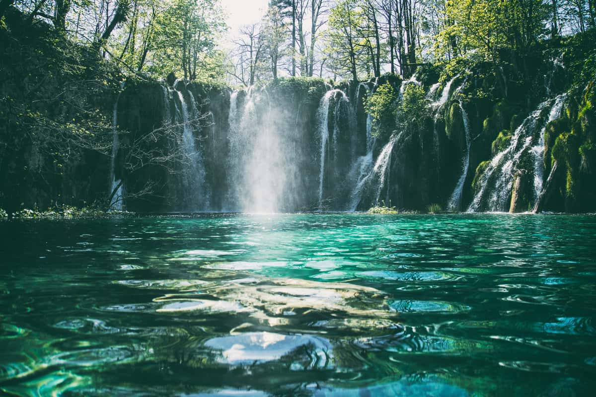 Scenic view of a turquoise pool with cascading waterfalls surrounded by lush, green forest under soft sunlight.