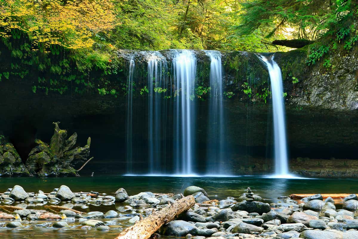 A serene forest waterfall pours in several narrow streams over a moss-covered cliff into a calm, rocky pool, surrounded by lush green and golden foliage.