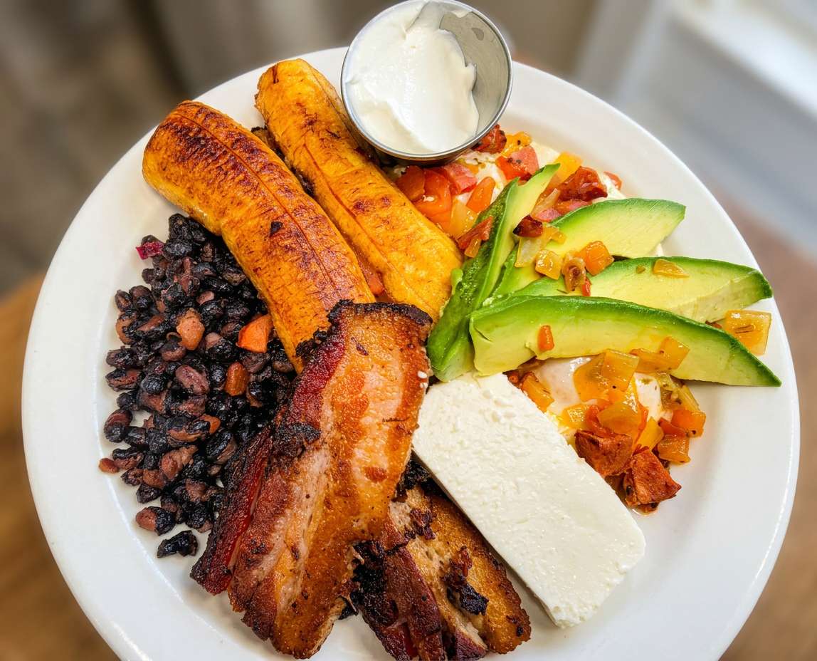 A plate with fried plantains, black beans, bacon, avocado slices, two eggs with salsa, white cheese, and a side of sour cream.
