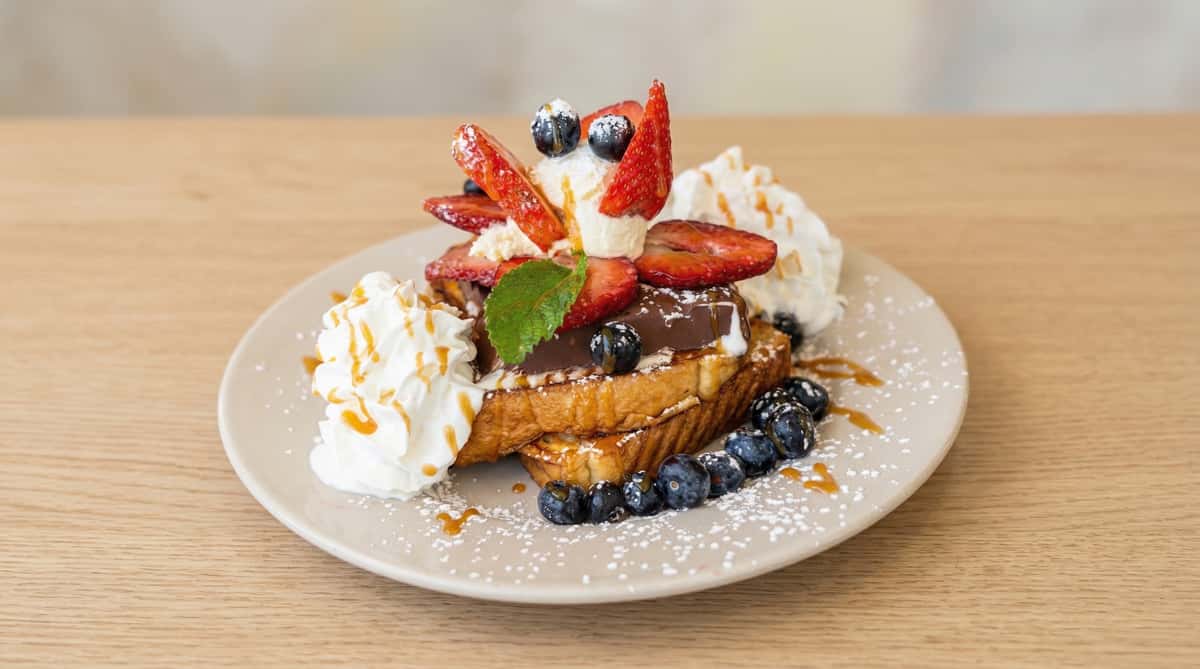 Decadent chocolate French toast topped with sliced strawberries, blueberries, whipped cream, caramel drizzle, and powdered sugar on a white plate.