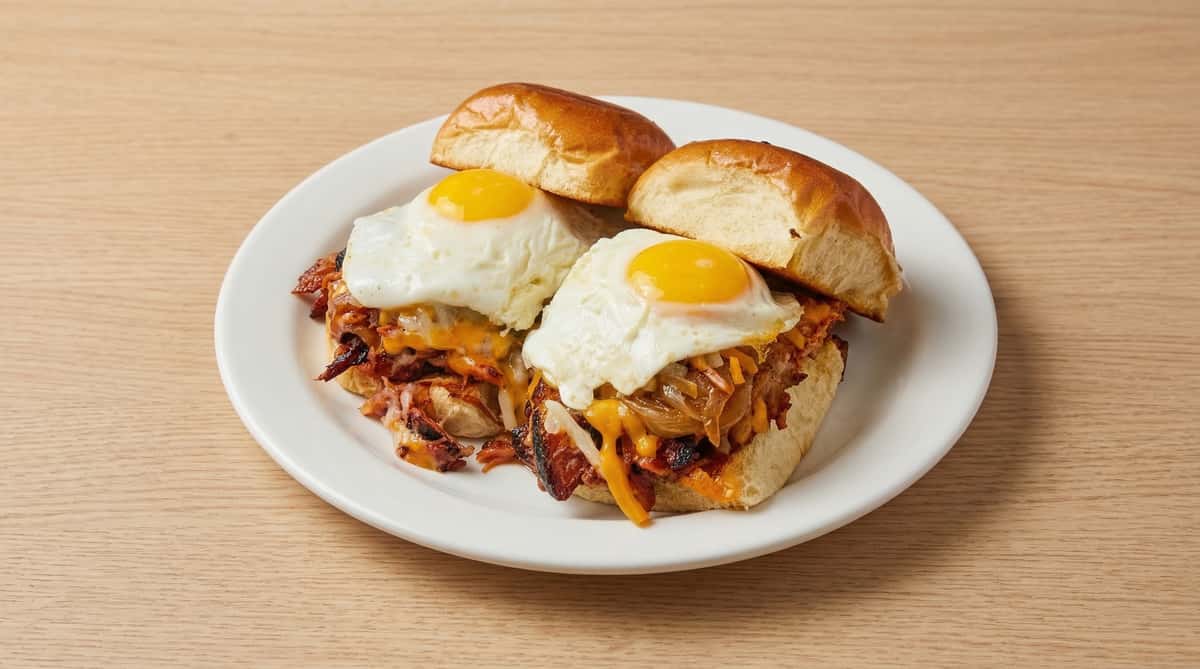 Breakfast sandwich with sunny-side-up eggs, melted cheddar, caramelized onions, and crispy bacon on a soft brioche bun, served on a white plate.