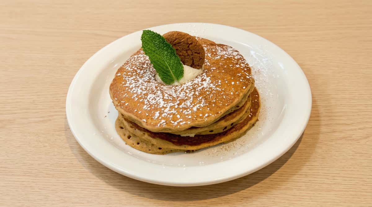 Fluffy stack of golden pancakes dusted with powdered sugar, topped with whipped butter, mint leaf, and a crisp cookie on a white plate in a cozy brunch setting.