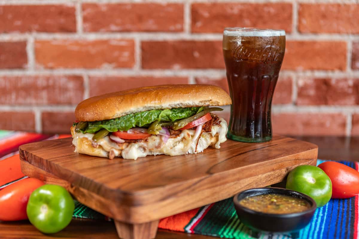 A Cencalli Taqueria cheesy torta sandwich with lettuce, tomato, and peppers, served with a soda on a wooden board atop a colorful Mexican serape against a brick wall background.