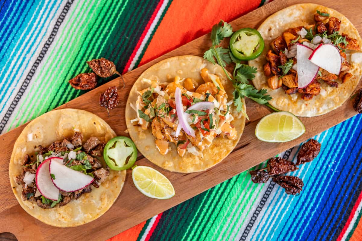 Three vibrant Cencalli Taqueria tacos with fresh toppings and lime wedges are presented on a wooden board against a colorful striped Mexican serape.