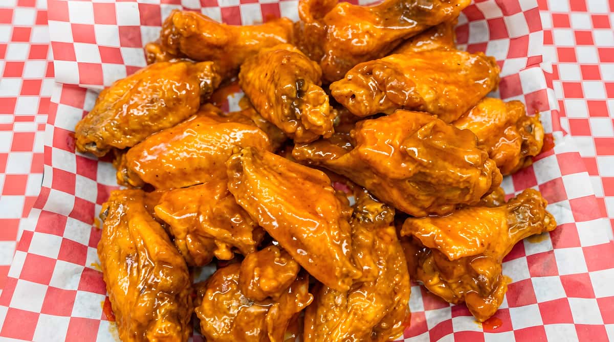 Pile of chicken wings coated in a shiny orange sauce, served in a basket lined with red and white checkered paper.