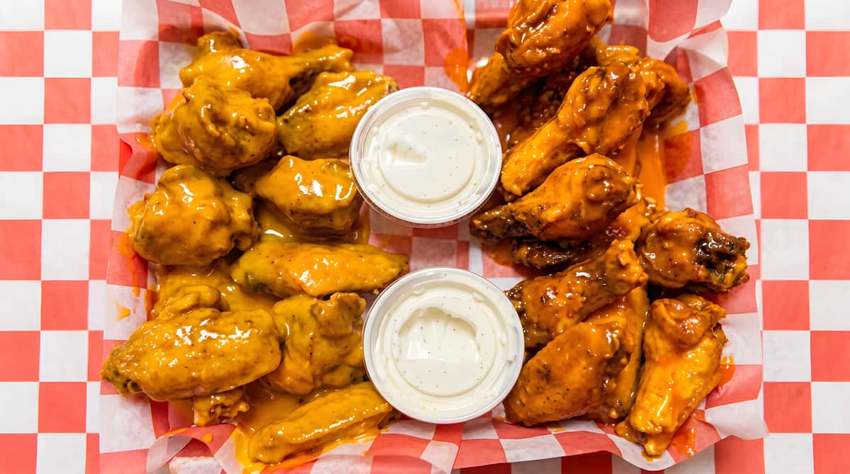 Two portions of chicken wings, some coated in orange sauce, others in reddish-orange sauce, served with white dipping sauces in paper baskets.
