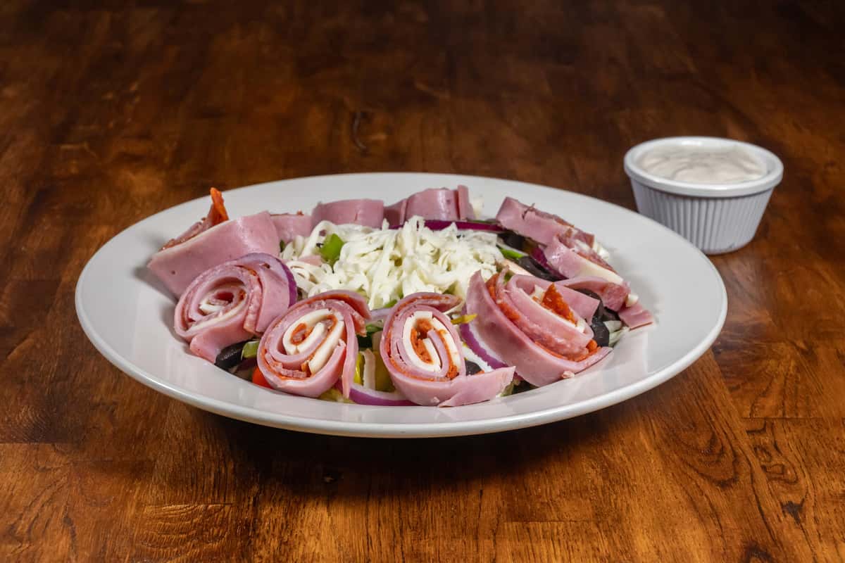 Italian antipasto salad featuring rolled ham, salami, and provolone over mixed greens with shredded mozzarella, red onions, olives, and bell peppers, served with a side of creamy dressing on a white plate.