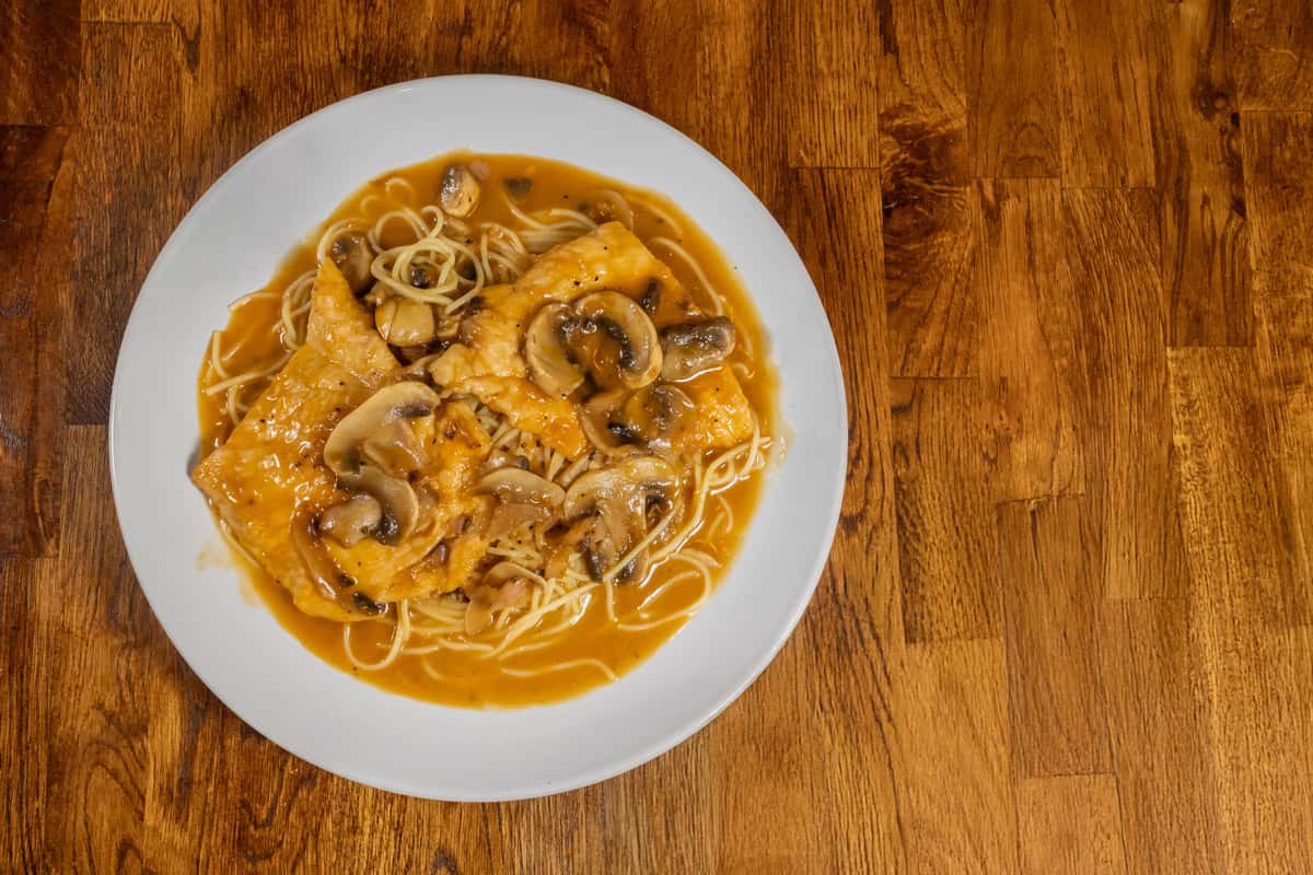 Chicken Marsala served over angel hair pasta, topped with sautéed mushrooms and smothered in a rich Marsala wine sauce on a white plate over a wooden table.