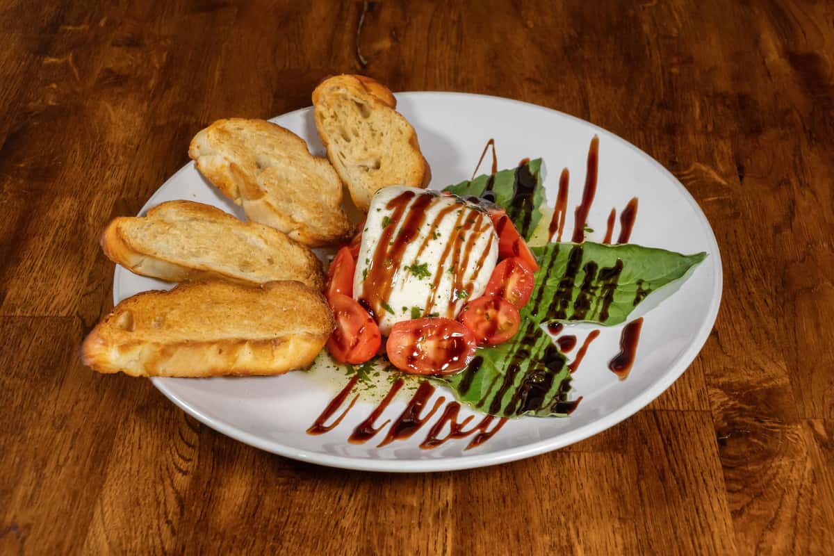 Fresh Caprese salad with creamy mozzarella, ripe cherry tomatoes, and basil leaves drizzled with balsamic glaze, served with toasted garlic crostini on a white plate over a rustic wooden table.