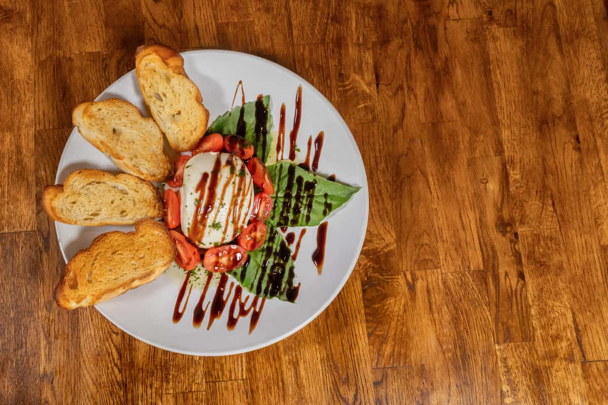 Caprese salad with fresh mozzarella, cherry tomatoes, and basil leaves drizzled in balsamic glaze, served with toasted crostini on a white plate over a wooden table.