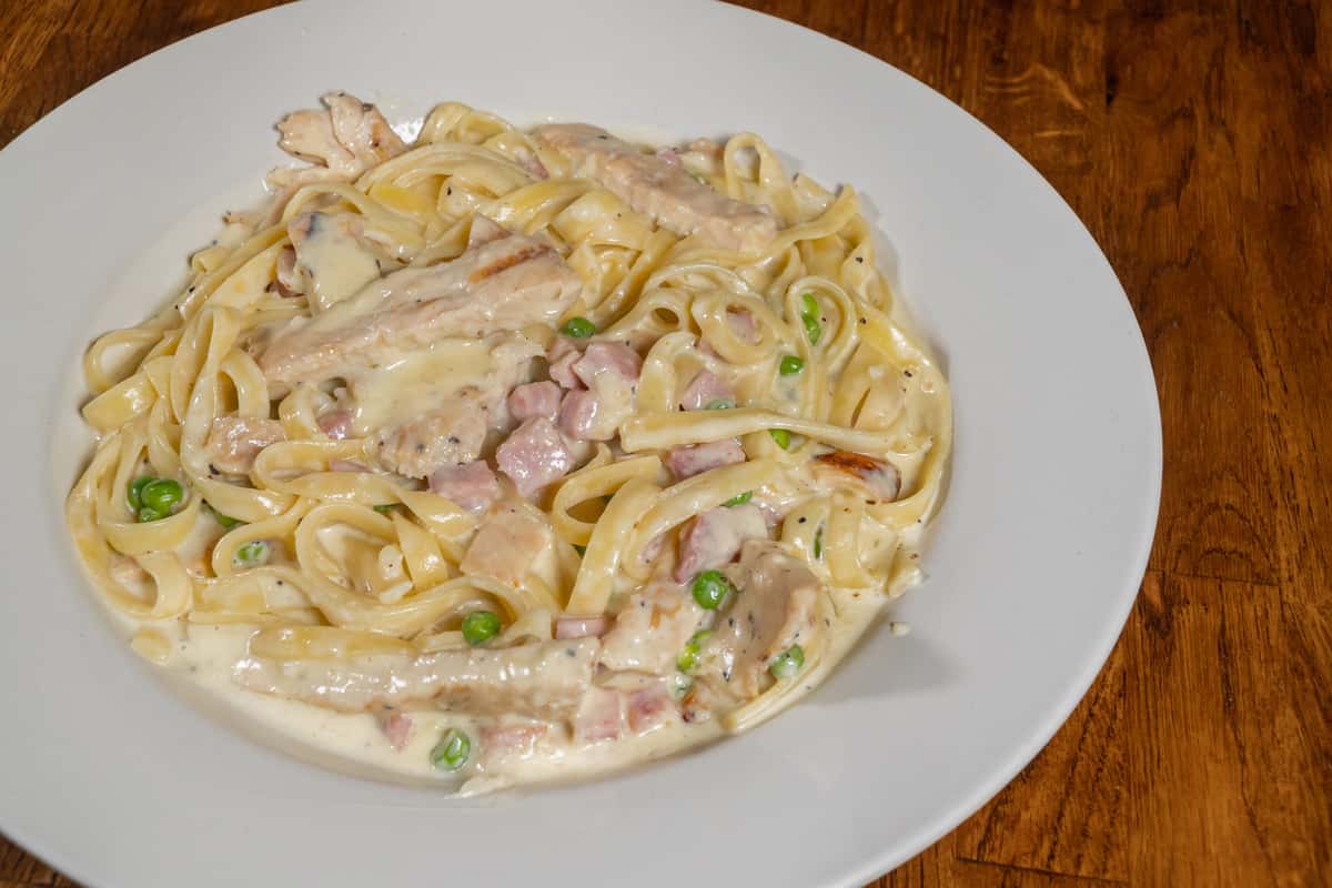Creamy fettuccine Alfredo tossed with grilled chicken, diced ham, green peas, and a rich garlic Parmesan sauce, served on a white plate over a wooden table.