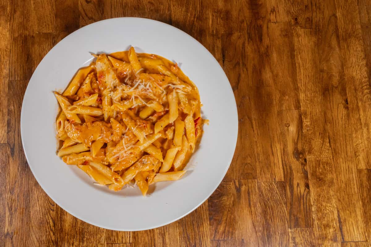 Creamy penne pasta with grilled chicken in a rich tomato cream sauce, topped with shredded Parmesan cheese and served on a white plate over a wooden table.