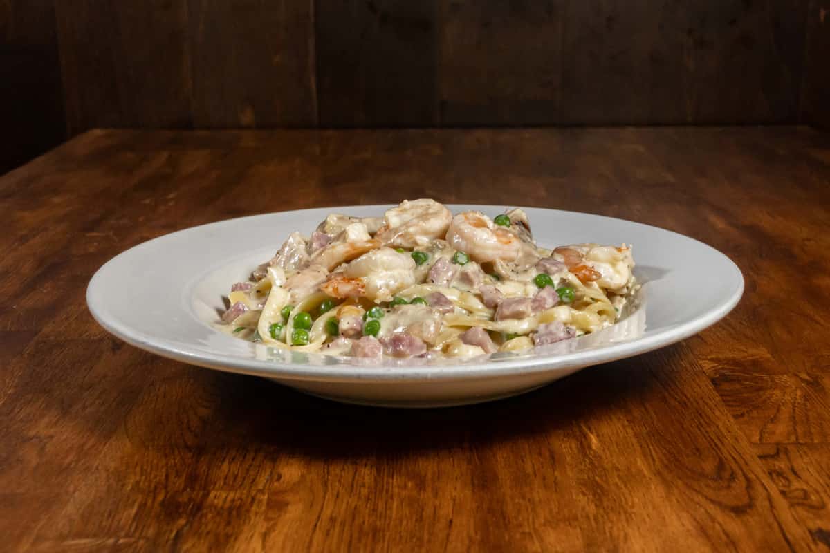 Creamy fettuccine pasta with shrimp, diced ham, and green peas in a rich Alfredo sauce, served on a white plate over a dark wooden table.