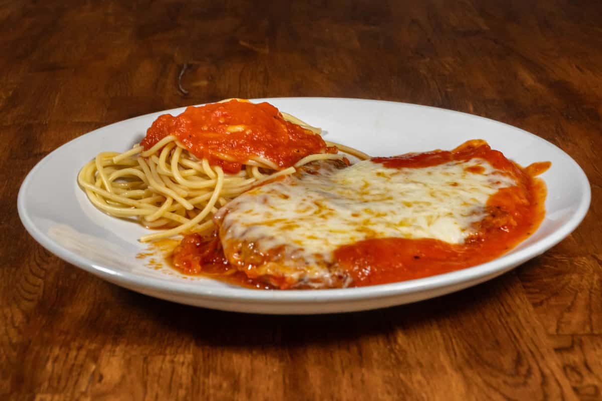 Classic chicken parmesan topped with melted mozzarella and marinara sauce, served alongside a portion of spaghetti with tomato sauce on a white plate over a rustic wooden table.