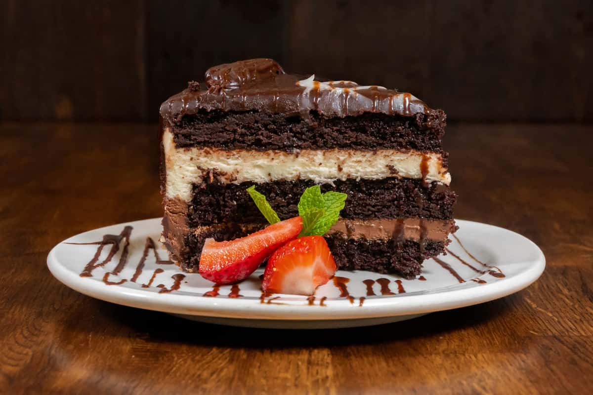 Decadent slice of layered chocolate cake with rich frosting and creamy filling, garnished with fresh strawberries and mint on a white plate with chocolate drizzle.