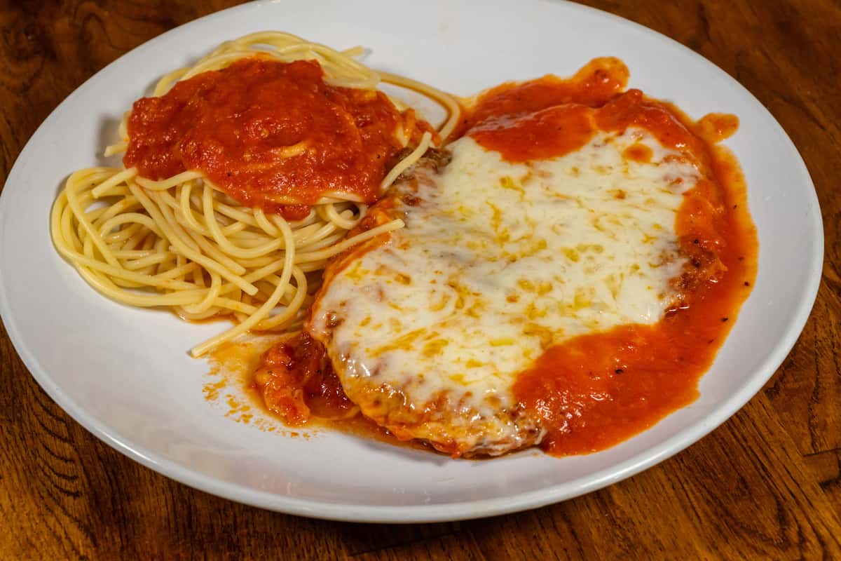 Classic chicken Parmesan topped with melted mozzarella and marinara sauce, served with a side of spaghetti in tomato sauce on a white plate over a wooden table.