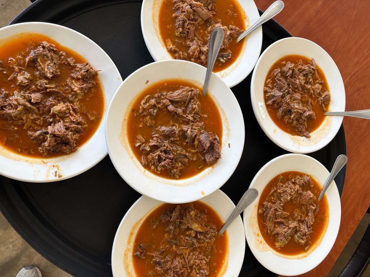 Six white bowls filled with tender birria beef in a rich red broth, each served with a metal spoon on a black tray.