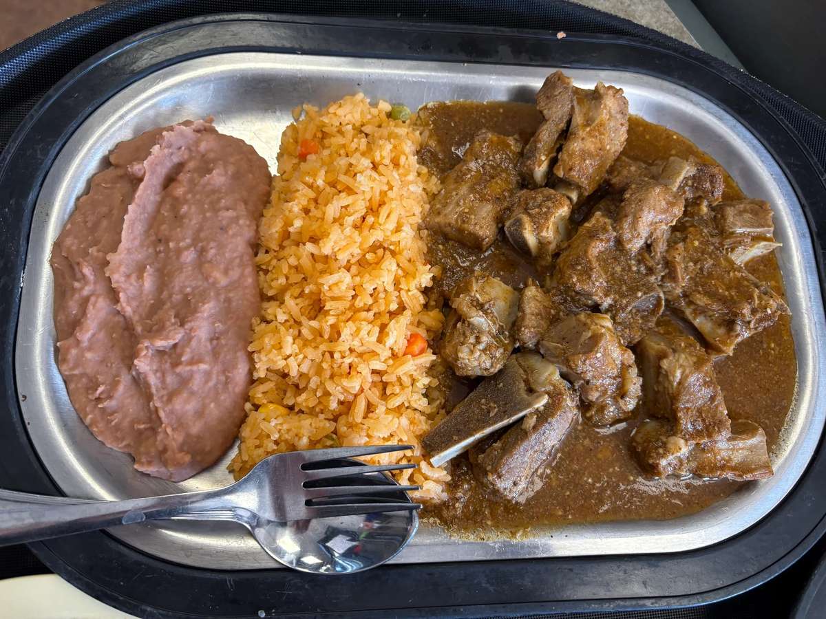 Mexican-style plate with refried beans, seasoned rice, and tender beef ribs in a rich brown sauce, served on a metal tray.