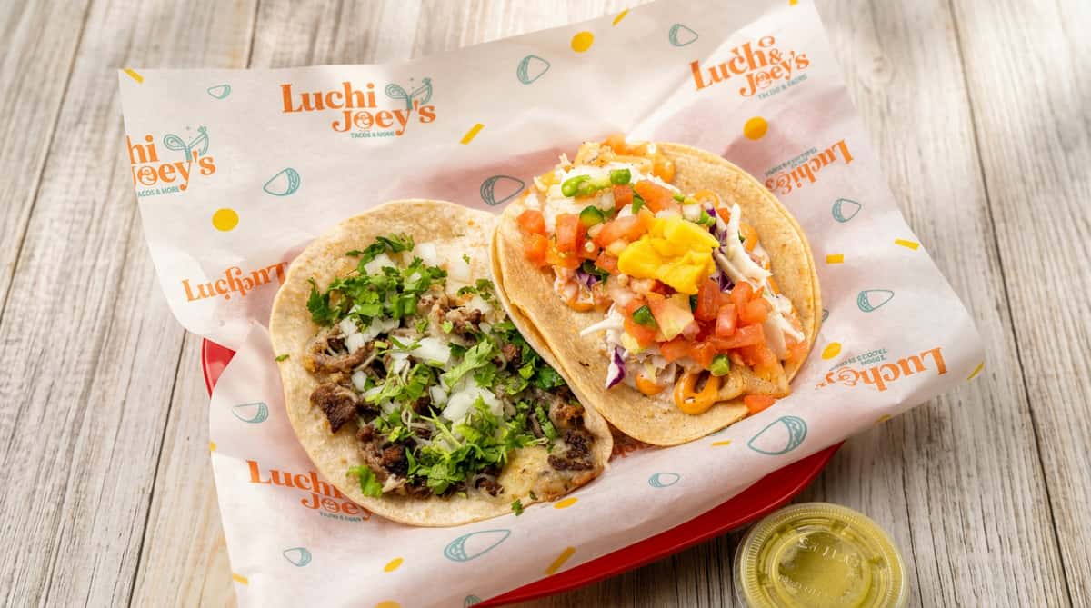 Two tacos on branded paper: one with meat, onions, cilantro; the other with fish, cabbage, salsa, and sauce.