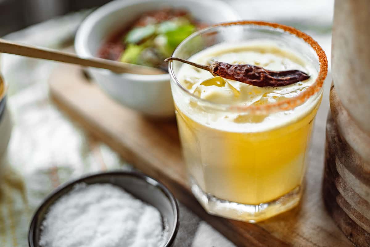 A refreshing spicy margarita garnished with a dried chili, featuring a rim of chili salt and a bowl of guacamole.
