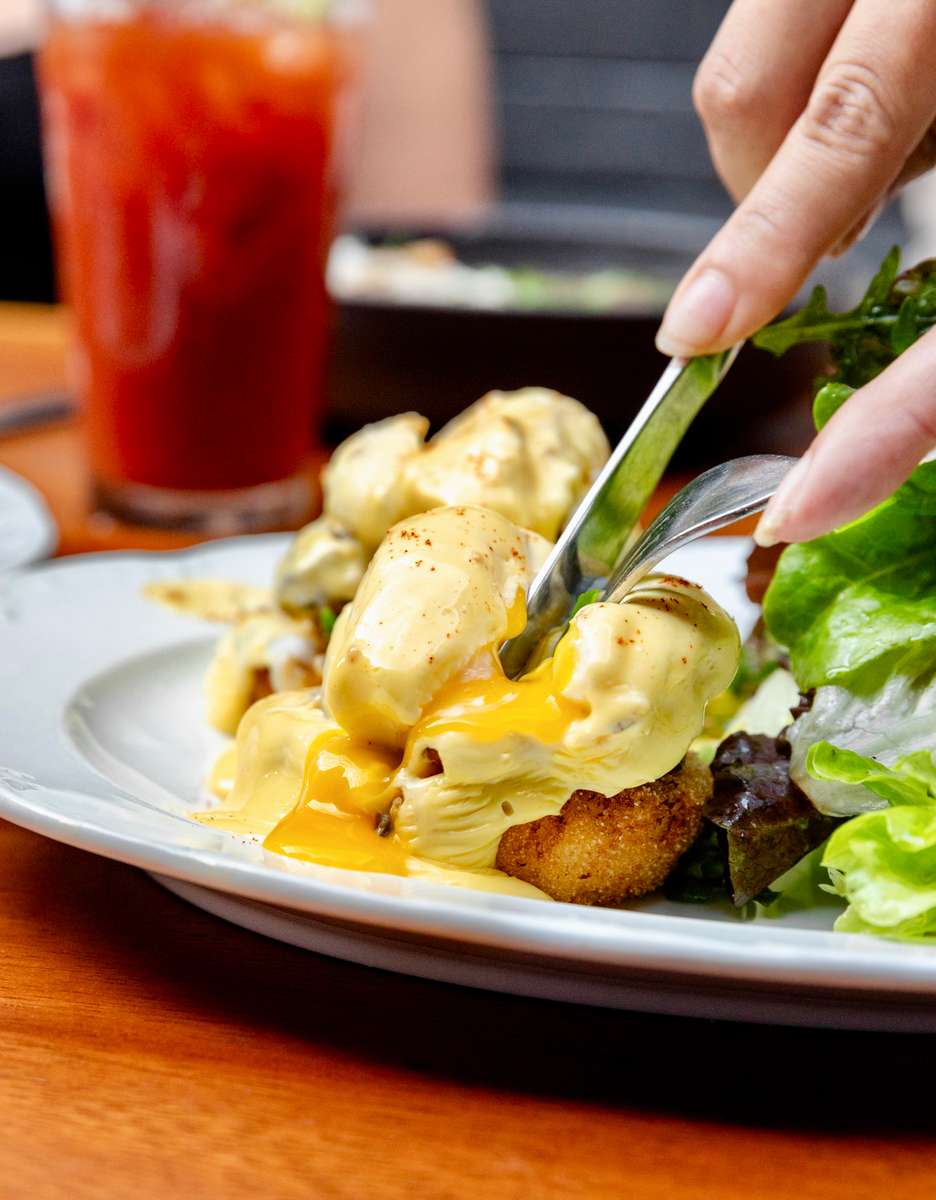 A rich and savory dish topped with perfectly poached eggs and creamy hollandaise sauce, gently cut open to reveal the golden yolk flowing over seasoned potatoes and greens, served with a refreshing iced drink for a balanced, satisfying meal.