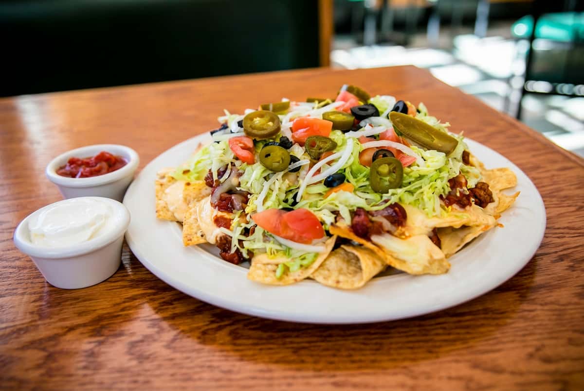 Loaded nachos with ground beef, cheese, shredded lettuce, tomatoes, onions, olives, and jalapeños. Salsa and sour cream are in small bowls.