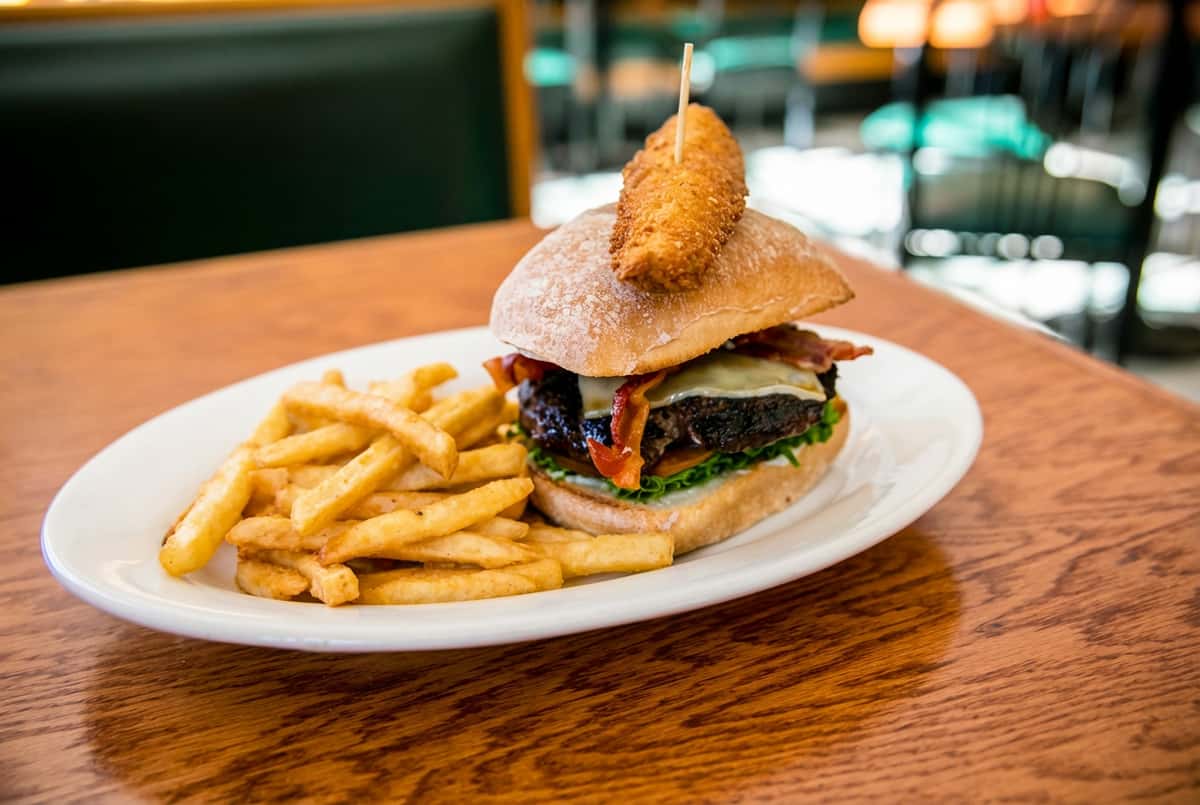 A burger with bacon, cheese, lettuce, and a fried chicken strip on top, served with a side of french fries.