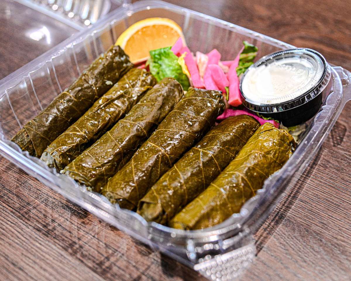 Stuffed grape leaves in a takeout container with pickled turnips, lettuce, an orange slice, and a side of garlic sauce