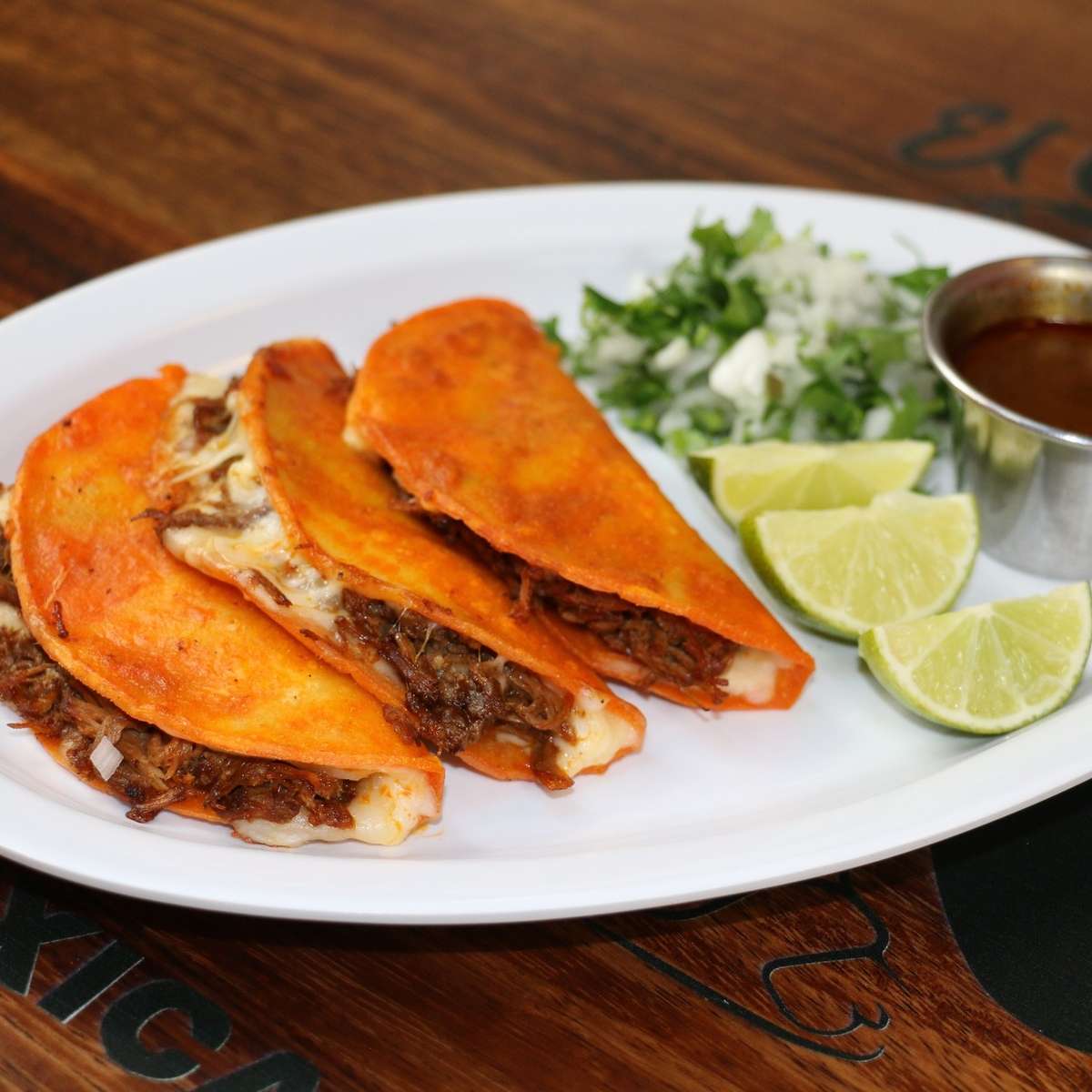 Three birria tacos with melted cheese, served with lime wedges, chopped onions and cilantro, and a side of consommé.