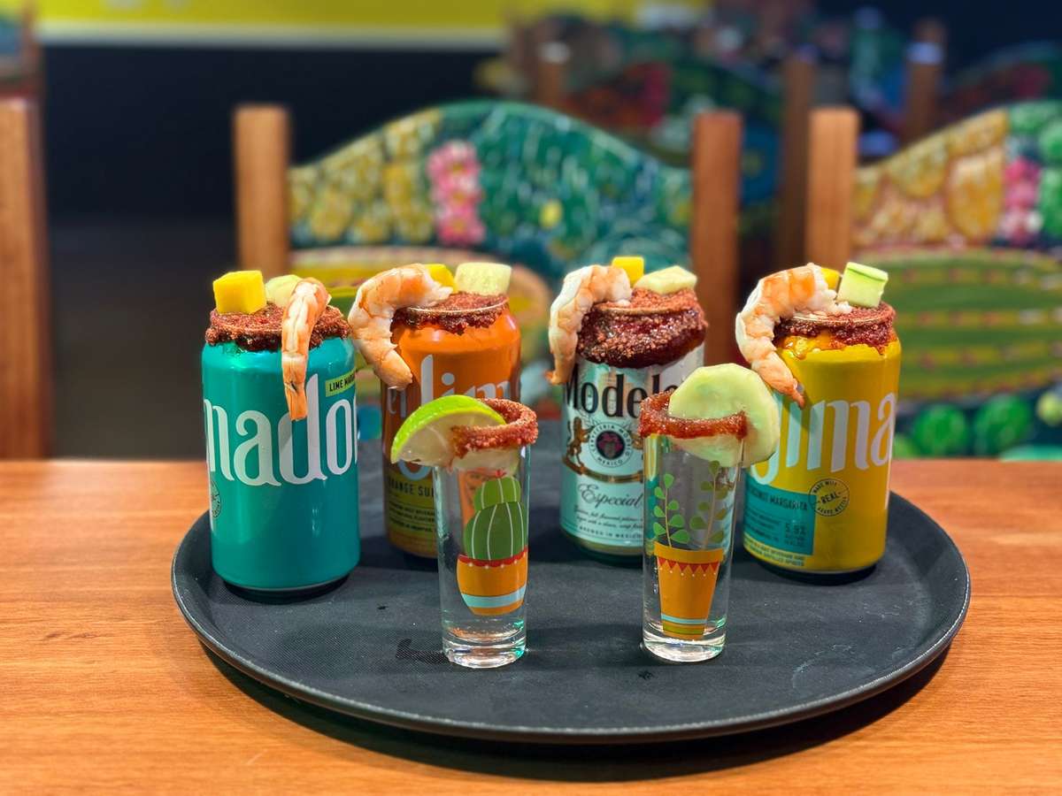 Assorted rimmed beer cans and shots topped with shrimp, lime, cucumber, and fruit, served on a black tray with colorful chairs.
