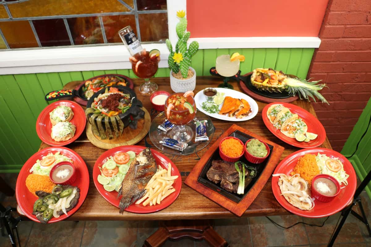 A vibrant spread of Mexican dishes and drinks on a wooden table, featuring seafood, tacos, grilled meats, and colorful cocktails.